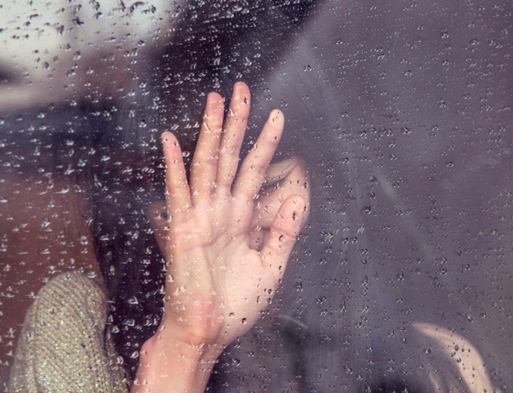 Photo d'une femme avec la main sur une vitre pleine de gouttes de pluie.